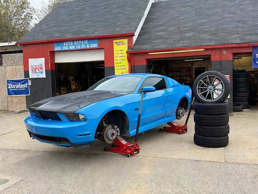 Mustang azul elevado con gatos rojos y cuatro llantas nuevas apiladas frente a MM Auto Tire Shop